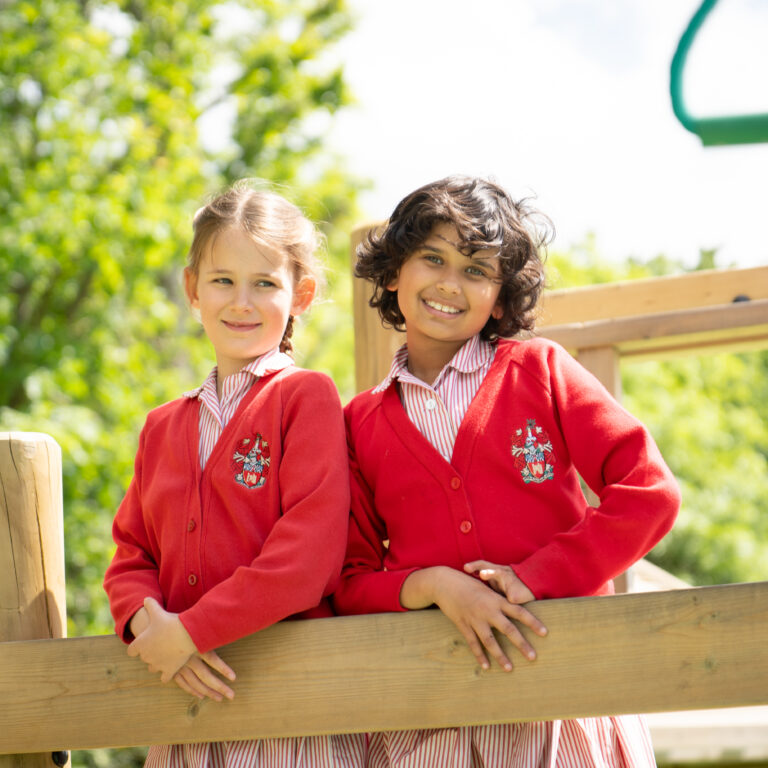 Two smiling children in red school uniforms lean on a wooden playground railing on a sunny day