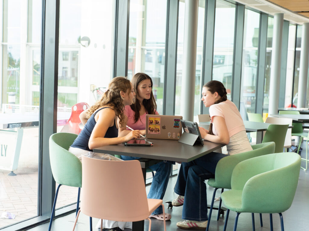 Three young people collaborate at a table with a laptop in a bright glass-walled study space. One points at the screen while another types, with a tablet nearby and snacks in view .