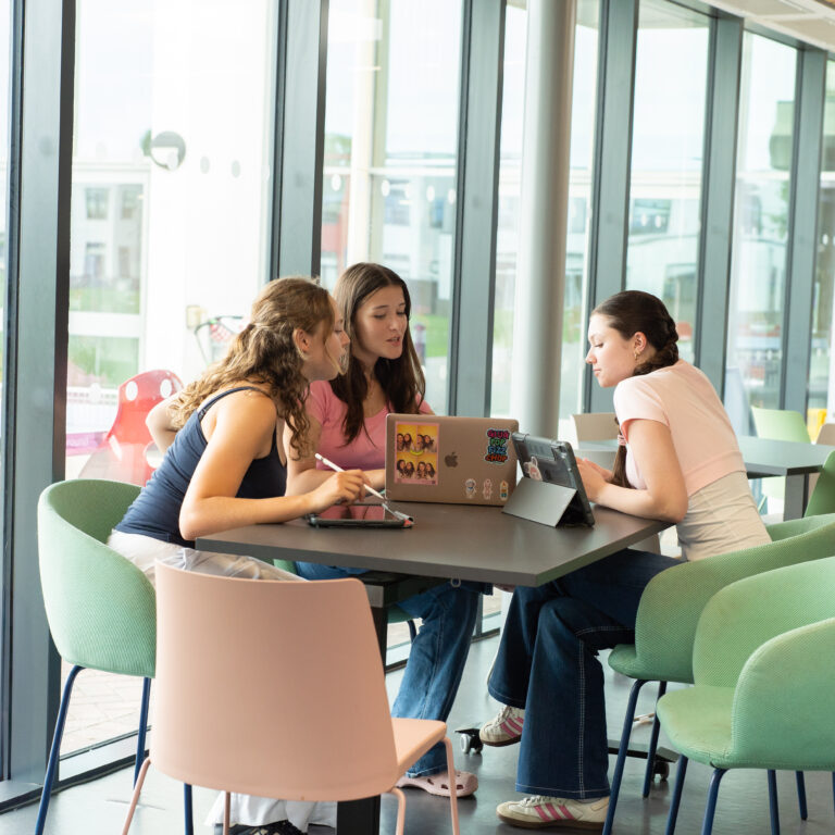 Three young people collaborate at a table with a laptop in a bright glass-walled study space. One points at the screen while another types, with a tablet nearby and snacks in view .