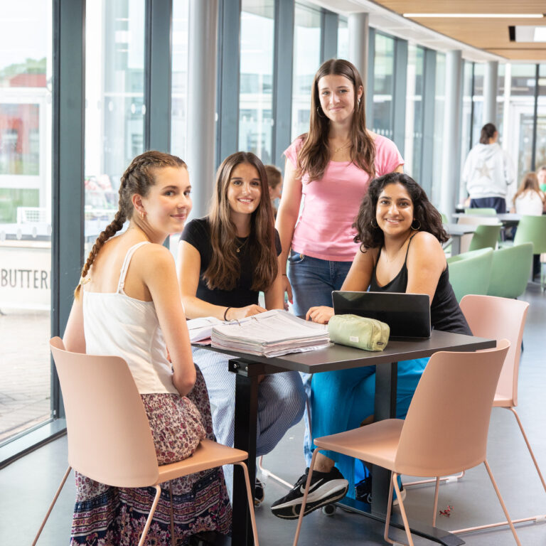 Four smiling students sit around a table with papers and a laptop in a bright glass-walled study area.
