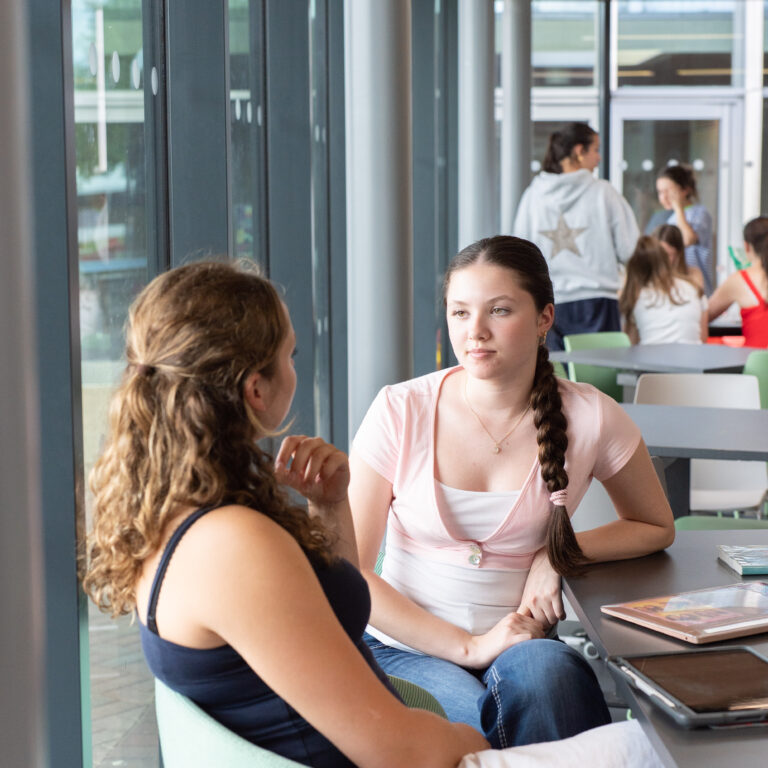 Two young women chatting at a table by large windows in a modern cafe setting.