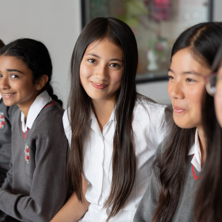 Three schoolgirls in uniform sit together and smile, with long dark hair and plaid skirts, indoors in a bright room.