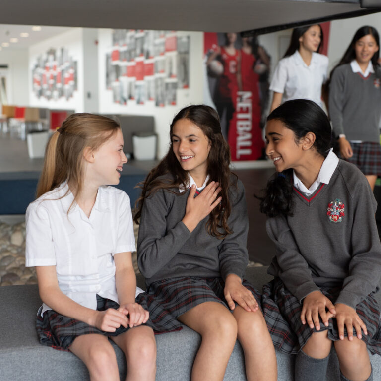 Group of five students in school uniforms relaxes in a modern common area, sitting and chatting on a gray bench.