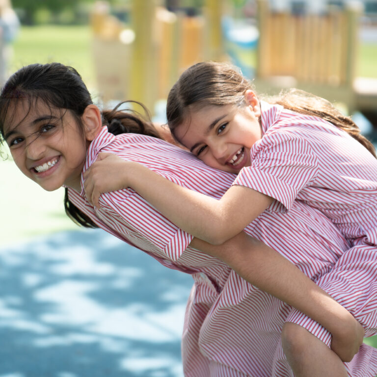 Two girls in red-and-white striped shirts share a joyful hug at a sunny playground.
