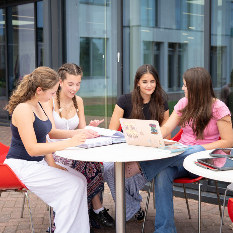 Group of five young women studying together at an outdoor table with notebooks and laptops.