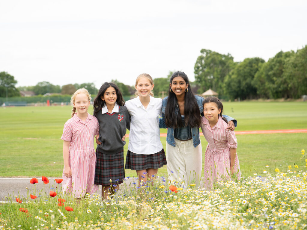 Five girls of diverse backgrounds stand arm in arm in a grassy field, smiling at the camera with a colorful flower border in front.
