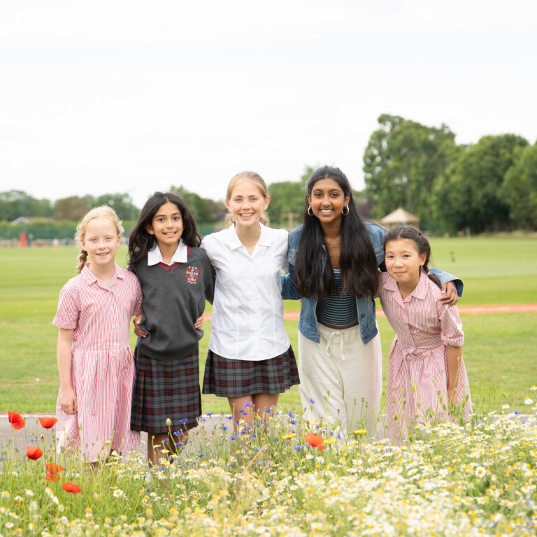 Five girls of diverse backgrounds stand arm in arm in a grassy field, smiling at the camera with a colorful flower border in front.