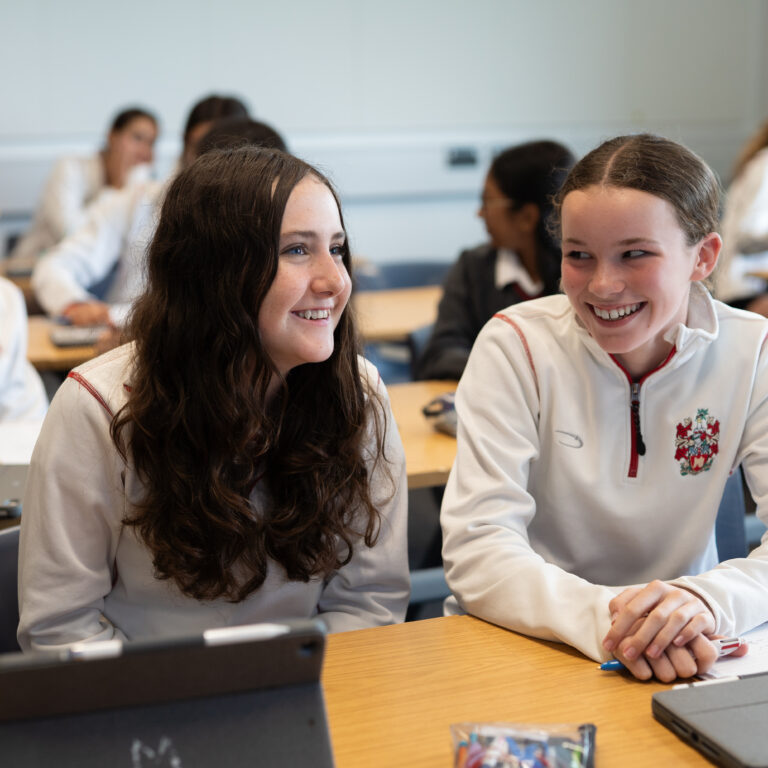 Two smiling students in white school uniforms sit at a desk in a classroom, with others chatting in the background.