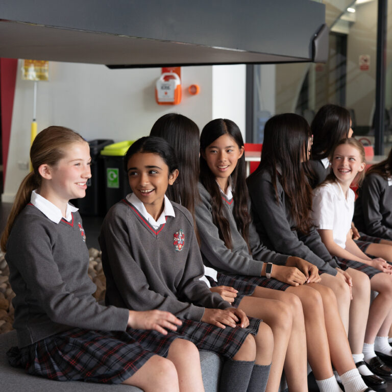 Group of smiling schoolgirls in gray sweaters and plaid skirts sit on a bench in a modern school lobby.