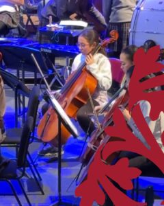 Young cellist wearing glasses and a light hoodie plays a cello in an orchestra rehearsal, with sheet music stands and other musicians nearby on a blue-lit stage.
