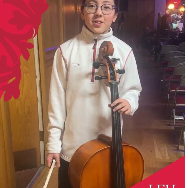 Young girl with glasses stands backstage, holding a cello and bow with stage seating in the background.