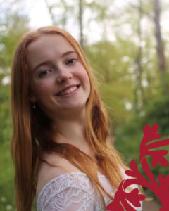 Smiling young woman with long red hair in a white lace top, outdoors among green trees; red botanical graphic in the bottom-right corner.