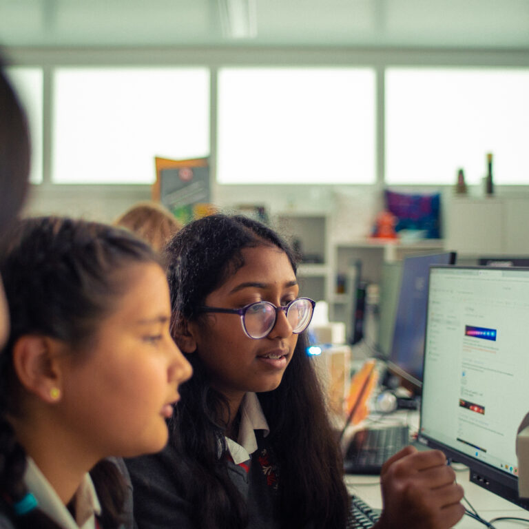 Three students in a bright computer lab focus on a monitor at a shared workstation, one foregrounded wearing glasses.