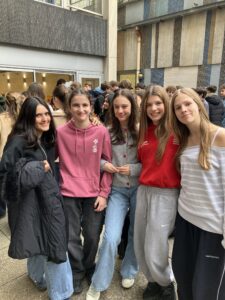 Five teenagers stand close together outdoors, smiling for a group photo in a school courtyard setting with a tiled building backdrop.