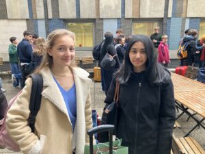 Two girls in coats stand in a schoolyard, smiling at the camera, with classmates and bags visible behind them on benches and near suitcases.