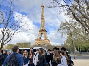 Group of schoolchildren with backpacks gathered in front of the Eiffel Tower in Paris on a cloudy day to pose for a photo.