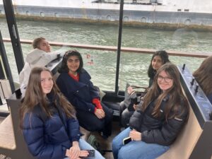 Six teens sitting in a glass-walled boat cabin, smiling, wearing jackets, with phones in hand and a river view outside the windows.