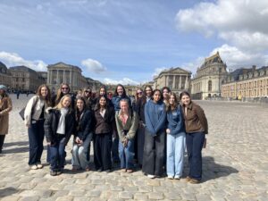 Group of young people smiling and posing on a cobblestone square in front of grand historic buildings under a blue sky.