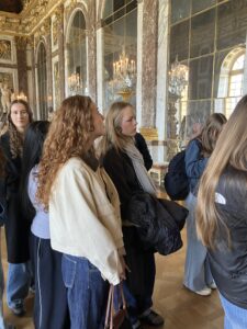 A tour group of young adults standing in a lavish marble-and-gold room with tall mirrors and crystal chandeliers, listening to a guide or display.