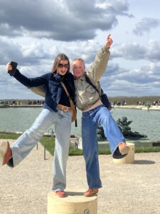 Two friends pose and jump on a stone pedestal by a fountain, one holding a phone for a selfie, with a cloudy sky and lake in the background.