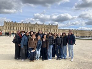 Group of students posing for a photo in front of the Palace of Versailles with a cloudy sky above, smiling at the camera.