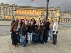 Group of young people posing together outdoors in front of a grand neoclassical building with illuminated windows and ornate street lamps at dusk/city square in Europe.
