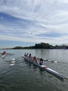 Crew team rowing in a long racing shell on a calm lake with spectators on the shore.