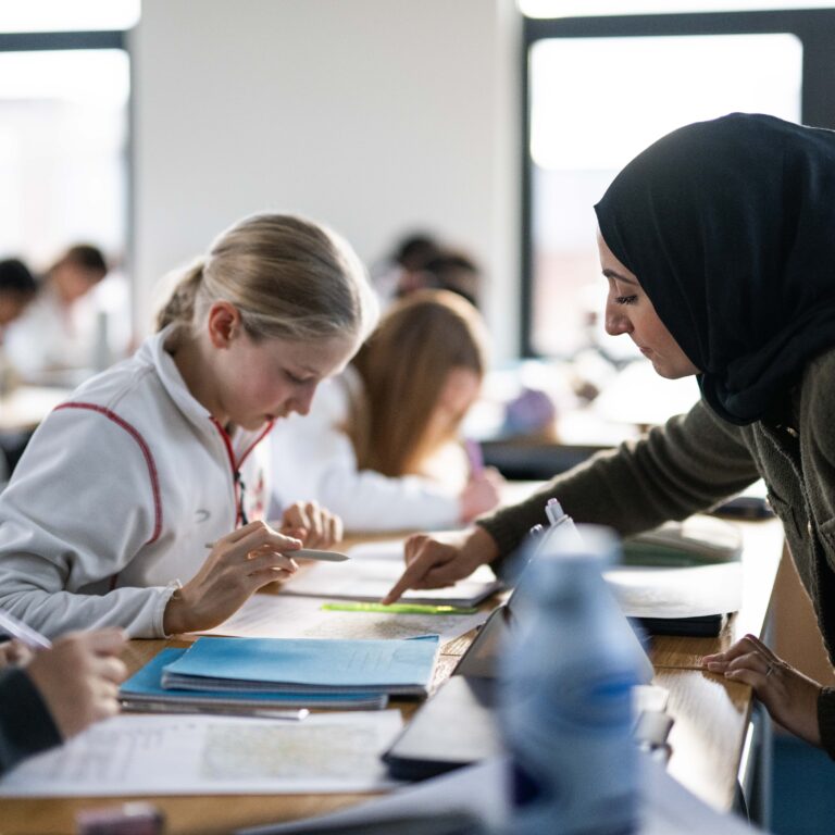 Teacher in a dark hijab leans over a desk to help students with worksheets in a bright classroom.