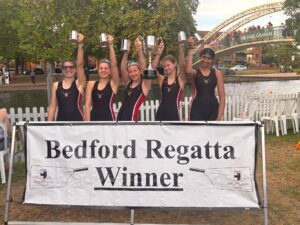 Five young female rowers in matching black uniforms cheer and raise multiple trophies in the air, standing behind a large Bedford Regatta Winners banner by the river.