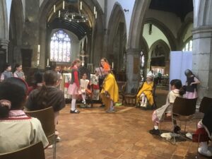 Children in period costumes perform a scene in a Gothic church nave, with onlookers seated along the sides and stone arches overhead.