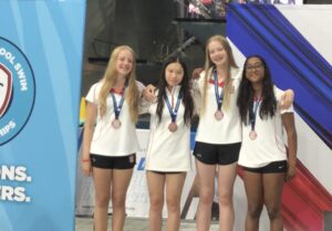 Four young female swimmers in white uniforms with red trim, wearing bronze medals, stand together and smile in front of a blue event backdrop with banners.
