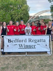 Group of young rowers in red shirts pose with trophies in front of a banner reading 'Bedford Regatta Winner' and a metal arch bridge in the background.