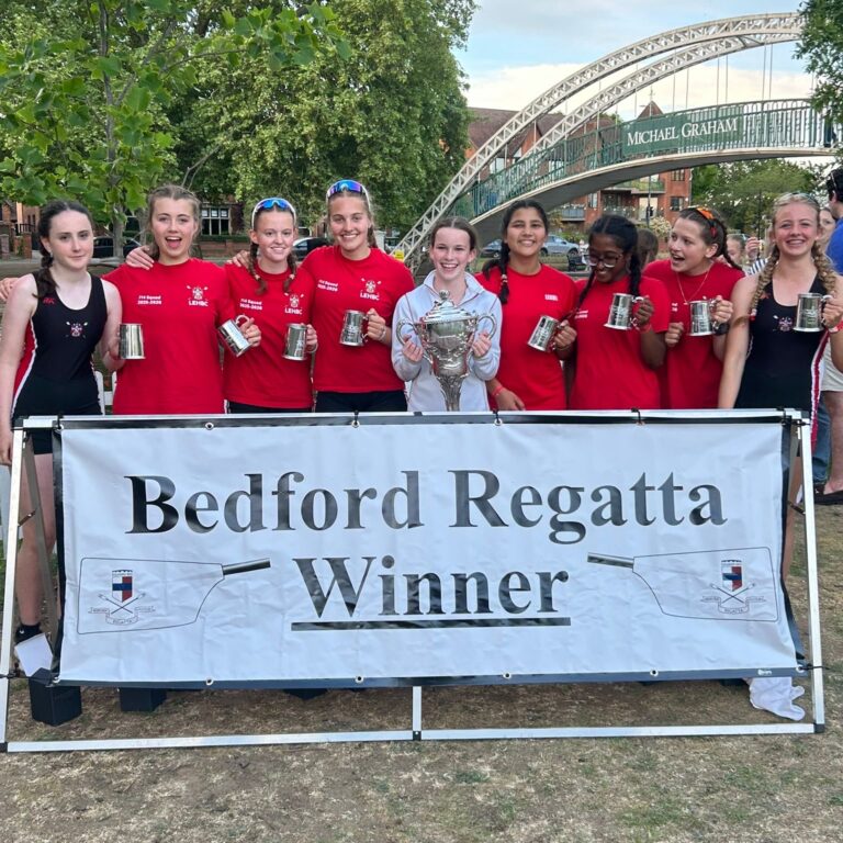 Group of young rowers in red shirts pose with trophies in front of a banner reading 'Bedford Regatta Winner' and a metal arch bridge in the background.