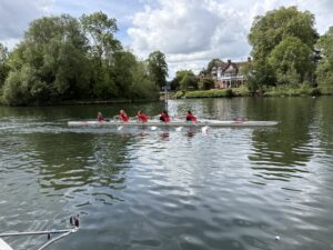 Rowing crew in red uniforms paddles a long boat on a calm river, with a brick manor and green trees along the bank in the background.