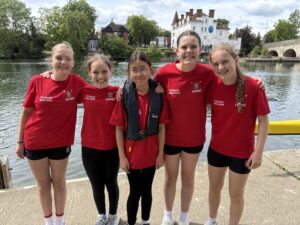 Five girls in red matching shirts stand arm-in-arm on a riverside walkway, smiling at the camera with a calm river and historic buildings in the background.