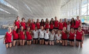 Group of young gymnasts in red tank tops and white shirts posing together in an indoor sports facility.