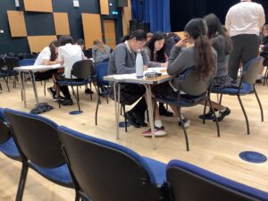 Students in uniform sit at tables, writing on worksheets in a large room with blue chairs and wooden wall panels.",