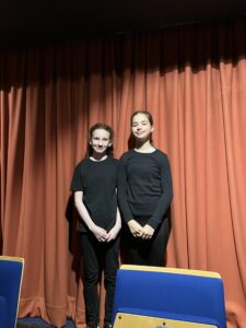 Two girls stand side by side on a stage in front of red curtains, wearing black outfits and smiling at the camera.