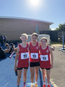 Three young female runners in red uniforms pose together on a blue track after a race, with bib numbers showing 19 and a sunny scene behind them.