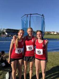 Three young female athletes in red uniforms with bib numbers 19 stand together on a blue running track, smiling with a stadium behind them.