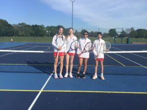Four young tennis players in white jackets and red skirts pose with racquets on a sunny outdoor court.