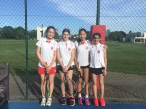 Four young girls in white tennis uniforms pose with rackets on an outdoor court behind a chain-link fence.
