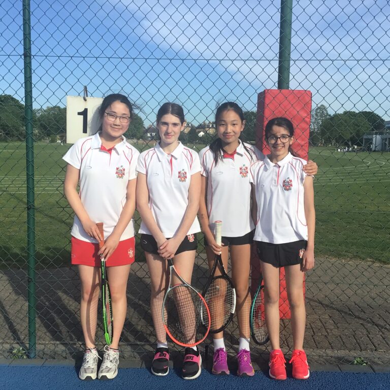 Four young girls in white tennis uniforms pose with rackets on an outdoor court behind a chain-link fence.