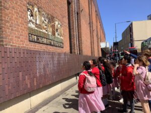 Group of children in red school uniforms standing along a brick wall with a large mosaic panel on the left side.