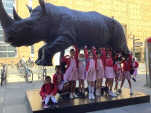 Group of schoolchildren in pink uniforms posing under a large black rhinoceros statue outdoors.