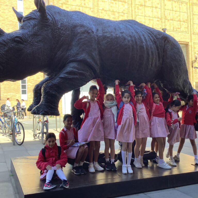 Group of schoolchildren in pink uniforms posing under a large black rhinoceros statue outdoors.