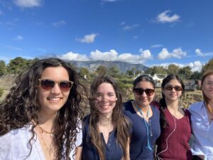 Five smiling women posing for a group selfie outdoors with mountains and a blue sky in the background