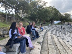 Group of young people sitting on concrete amphitheater steps outdoors, chatting with a rocky hillside and trees behind them.