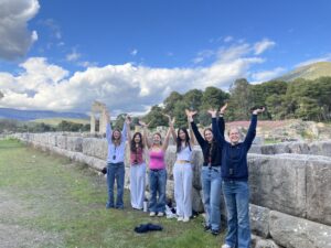 Group of young people standing in front of ancient stone ruins with their arms raised in the air, smiling at the camera, under a blue sky with scattered clouds and hills in the background.