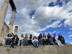 Group of about 15 students sitting on ancient stone steps, with weathered pillars on the left and a blue, cloudy sky above.